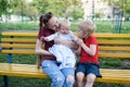 Young mother and two children on a bench in the park. Baby and blond elder brother playing. Spring warm day