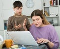 Mother working at laptop and unhappy preteen son standing near table