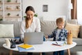 Young mother working online on laptop with son playing around, lady freelancing and taking care about her toddler child Royalty Free Stock Photo