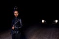 A young  mulatto girl in a leather jacket and black clothes poses on an abandoned sand road. at night in the light of car Royalty Free Stock Photo