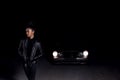 A young  mulatto girl in a leather jacket and black clothes poses on an abandoned sand road. at night in the light of car Royalty Free Stock Photo