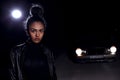 A young  mulatto girl in a leather jacket and black clothes poses on an abandoned sand road. at night in the light of car Royalty Free Stock Photo