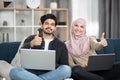 Young multiracial Muslim couple relaxing on sofa at home using laptops, smiling at camera and showing thumbs up Royalty Free Stock Photo