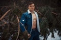 Young muscular man in unbuttoned jacket with bared breast stands next to pine tree in winter forest. Royalty Free Stock Photo