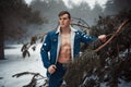 Young muscular man in unbuttoned jacket with bared breast stands next to pine tree in winter forest. Royalty Free Stock Photo