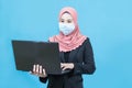 Young muslim asian business woman with traditional dress in black suit wearing medical face mask holding laptop isolated on blue Royalty Free Stock Photo