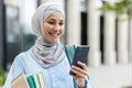 Young muslim woman in hijab walking outside university campus, female student smiling contentedly using app on phone Royalty Free Stock Photo