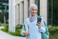 Young muslim woman in hijab walking outside university campus, female student smiling contentedly using app on phone Royalty Free Stock Photo