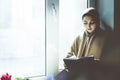 Young Muslim woman sits on the window sill. Middle-eastern writer woman sitting thoughtfully near the window and writing her story