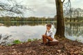 A young nipper guy in retro old-fashioned village clothes sits saddened on an old suitcase propped his head on the bank of a river