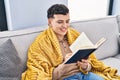 Young non binary man reading book sitting on sofa at home Royalty Free Stock Photo