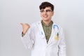 Young non binary man wearing doctor uniform and stethoscope smiling with happy face looking and pointing to the side with thumb up Royalty Free Stock Photo