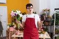 Young non binary man working at florist shop showing smartphone screen in shock face, looking skeptical and sarcastic, surprised