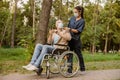 Young nurse in face shield and protective face mask assisting senior handicapped man in wheelchair using his smartphone Royalty Free Stock Photo