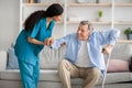 Young nurse helping elderly man with crutches to get up from sofa indoors Royalty Free Stock Photo