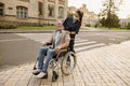 Young nurse in protective face mask taking care of senior handicapped man in wheelchair during a walk in the city Royalty Free Stock Photo