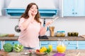 young overweight woman with wooden spoon in hand talking on smartphone while standing at table with fresh vegetables in kitchen Royalty Free Stock Photo