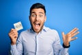 Young overworked business man asking for help holding paper over blue background very happy and excited, winner expression