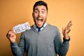 Young overworked business man asking for help holding paper over yellow background very happy and excited, winner expression