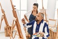 Young painting teacher woman teaching art to senior man painting on canvas at art studio