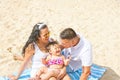Young Parents Cute Laughing Baby Toddler Daughter with Kiss Mark on Cheek Sitting on Beach Smiling Looking at Each Other. Summer