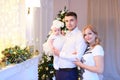 Young parents keeping little female baby near Christmas tree in background.