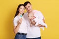 Young parents wearing white casual t shirts posing with new born daughter, mother showing toy for little baby with nippel,