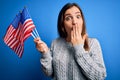 Young patriotic woman holding usa flag on independence day 4th of july over blue background cover mouth with hand shocked with