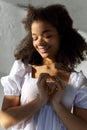 Young peaceful grateful thankful african american woman with closed eyes in white dress making heart sign, led lights in hands of