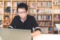 Young pensive Asian man working on laptop at home office or library with serious face, bookshelf blur background with copy space