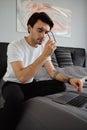 Young pensive brunette man in T-shirt sitting on big bed thoughtfully taking off eyeglasses while working on laptop at Royalty Free Stock Photo
