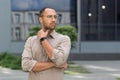 Young pensive businessman in glasses outside modern office building, man thinking and looking away, office worker in Royalty Free Stock Photo