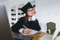 Young pensive caucasian woman college student in graduation outfit , sitting in front laptop with books and thinking about future Royalty Free Stock Photo