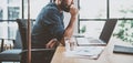 Young pensive coworker working at sunny work place loft while sitting at the wooden table.Man analyze document on laptop