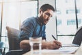 Young pensive man working at sunny loft office on laptop.Businessman making notes on paper documents reports.Blurred Royalty Free Stock Photo