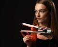 Young pensive woman with long silky straight hair in black bodysuit holds demonstrates hair straightener and looks aside