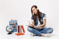 Young pensive woman student in headphones listening music writing notes on notebook sitting near globe backpack, school