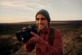 Young photographer with backpack and cap holding professional camera and shooting morning sunrise