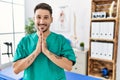Young physiotherapist man working at pain recovery clinic praying with hands together asking for forgiveness smiling confident Royalty Free Stock Photo