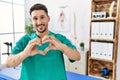 Young physiotherapist man working at pain recovery clinic smiling in love doing heart symbol shape with hands Royalty Free Stock Photo