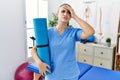 Young physiotherapist woman holding yoga mat stressed and frustrated with hand on head, surprised and angry face Royalty Free Stock Photo