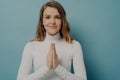 Young pleaded trustful woman posing with hands folded in praying gesture, isolated on blue