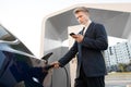 Young pleasant Caucasian businessman uses smart phone while waiting for charging the battery in his modern electric car