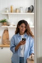Young pleasant smiling woman standing in kitchen holding smartphone, watching cooking video recipe