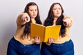 Young plus size twins holding book pointing with finger to the camera and to you, confident gesture looking serious Royalty Free Stock Photo