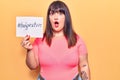 Young plus size woman holding paper with hashtag body positive scared and amazed with open mouth for surprise, disbelief face