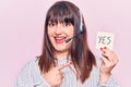 Young plus size woman wearing call center agent headset holding yes reminder smiling happy pointing with hand and finger