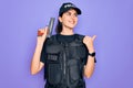 Young police woman wearing security bulletproof vest uniform and holding gun pointing and showing with thumb up to the side with Royalty Free Stock Photo