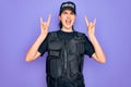 Young police woman wearing security bulletproof vest uniform over purple background shouting with crazy expression doing rock