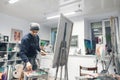 Young, positive artist works, paints a picture of oil paints in a cozy studio. Artist with a canvas on an easel places an oil Royalty Free Stock Photo